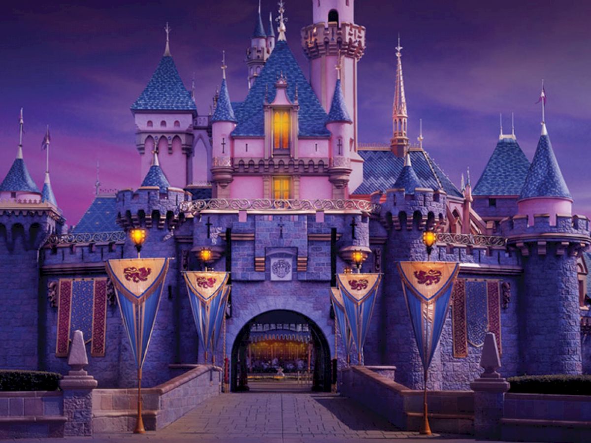 The image shows a beautifully lit-up fairy tale castle with blue roofs, flags, and warm glowing lights against a twilight sky.