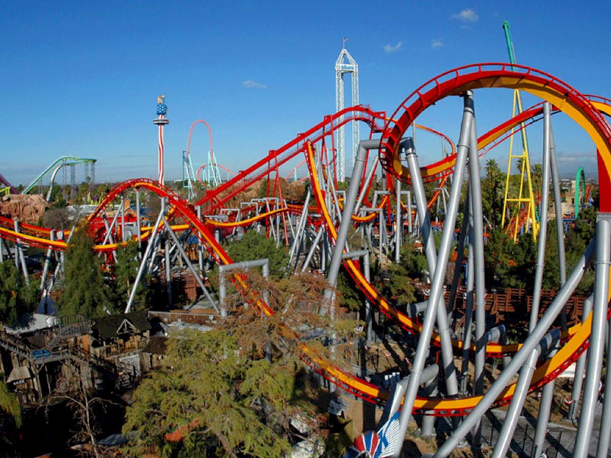 The image shows a large, colorful roller coaster with multiple loops and turns set within an amusement park, surrounded by trees, with a clear blue sky.