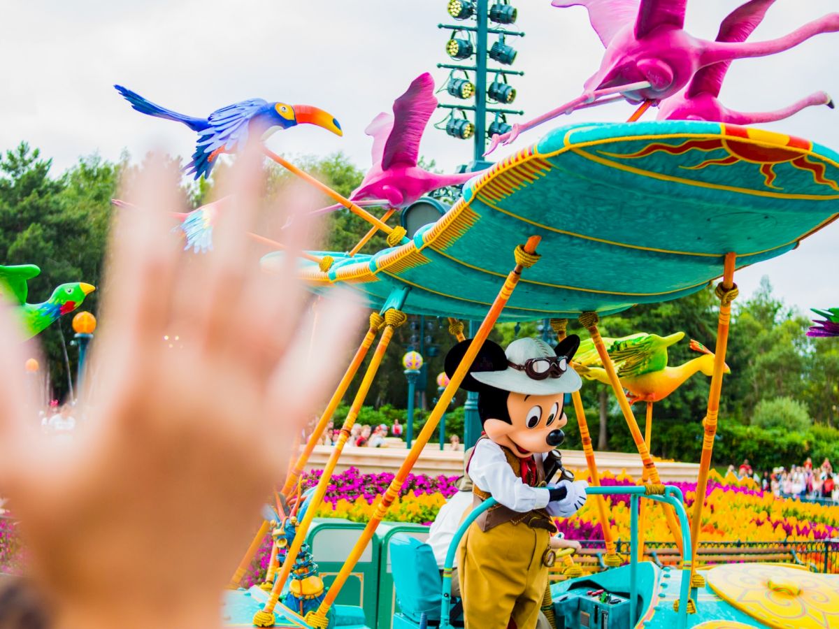 A person waves at a character in a Disney-themed parade, featuring colorful birds and vibrant decorations in the background.