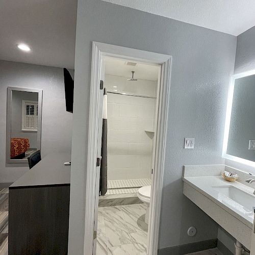 This image shows a bathroom with a shower, vanity with mirror, and a partial view of an adjacent area with a desk and chair in neutral tones, ending the sentence.