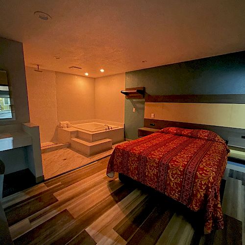 A hotel room with a double bed, colorful bedspread, wooden floor, and a jacuzzi bathtub in the corner, softly lit.