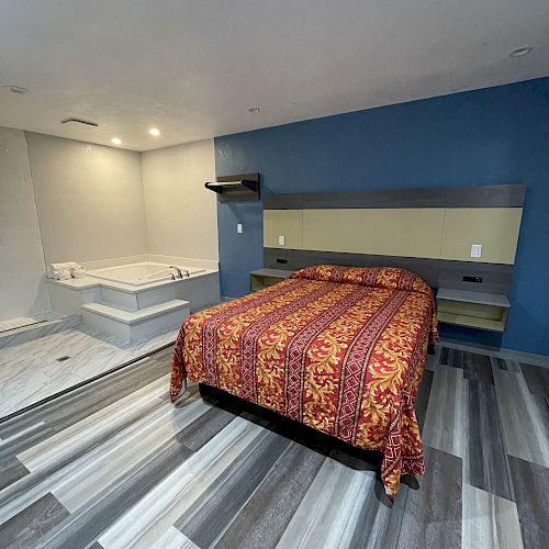 A modern bedroom with gray-striped flooring, a blue accent wall, a bed with a colorful bedspread, and a corner jacuzzi tub.