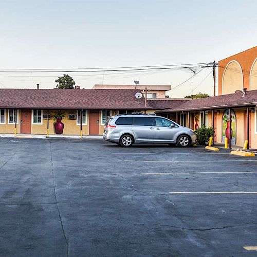 A parking lot in front of a motel with a gray van and additional empty parking spaces. The building is orange and yellow with decorative wall arches.