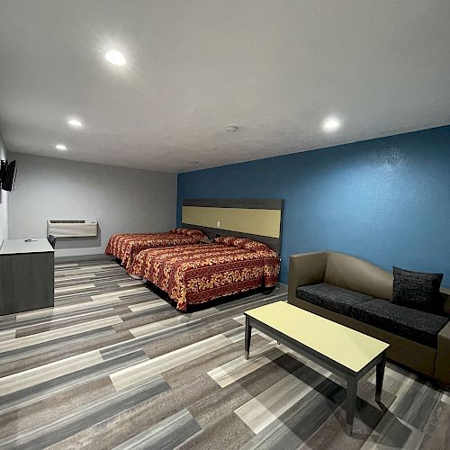 A room with two beds, a blue accent wall, a couch, a table, and a TV mounted on the wall. The flooring is a wood pattern.