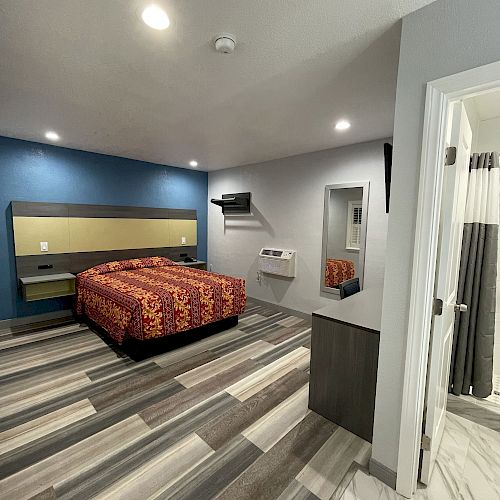 A modern bedroom with a bed having a colorful bedspread, a blue accent wall, striped flooring, and a bathroom visible in the corner ending the sentence.