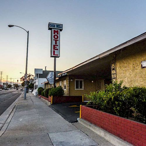 A street view with a "Motel" sign, sidewalk, street lamp, and shrubs beside a building at sunset.