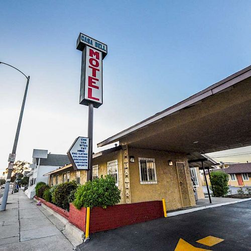 The image shows an exterior view of a motel with a large red and white sign reading "MOTEL." The building is situated next to a paved road.