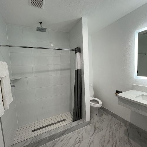 The image shows a modern bathroom with a walk-in shower, a floating sink, a backlit mirror, and a toilet on a gray marble floor, well-lit by overhead lights.