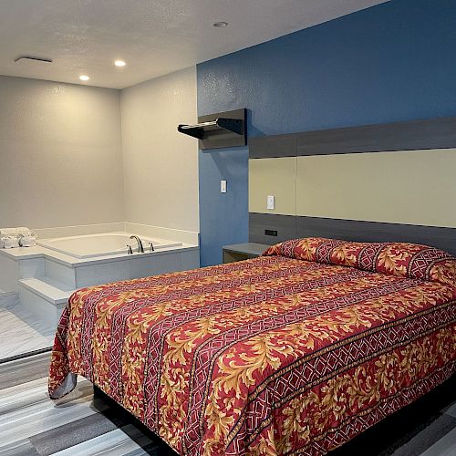 The image shows a hotel room with a bed covered in a colorful patterned bedspread and a jacuzzi bathtub in the corner.