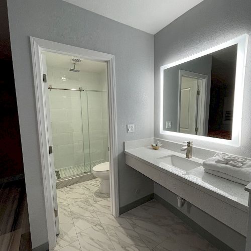 A modern bathroom with a large illuminated mirror, a sink with a countertop, and a separate shower area with a glass door visible through an open door.