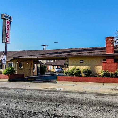 A single-story motel with a prominent sign is situated along a street, featuring a small entryway and well-maintained landscaping.