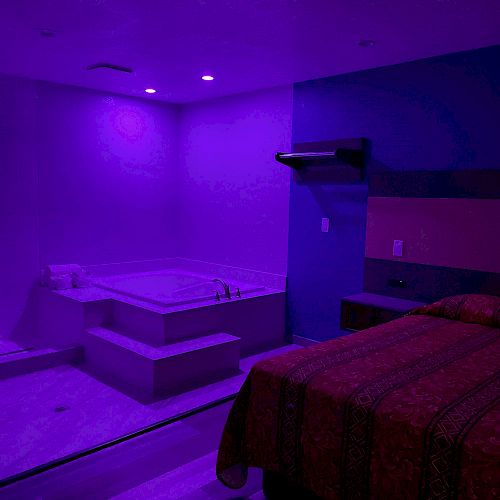 A bedroom with purple lighting, featuring a bed, a wall-mounted TV, a desk, and a built-in corner bathtub.