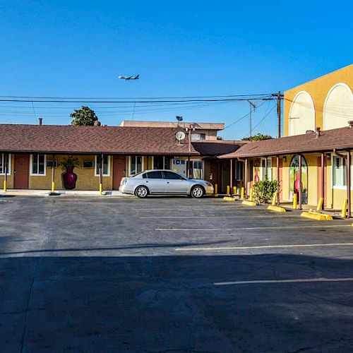 The image shows a motel with yellow walls and a shingled roof, featuring a central courtyard and parking area with two cars parked. An airplane is above.