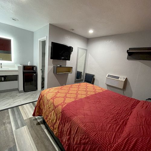 The image shows a small, modern bedroom with a bed, wall-mounted TV, shelf, air conditioning unit, and an open door leading to a bathroom with a sink.