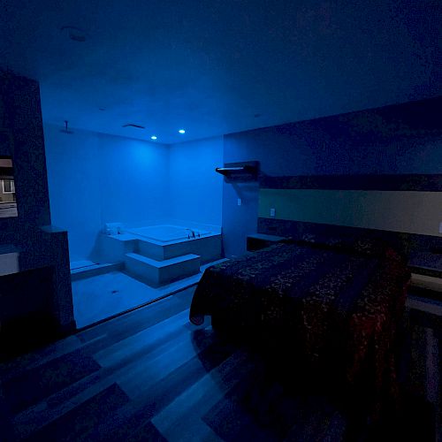 The image depicts a dimly lit room with blue lighting, featuring a bed and a bathtub in the corner. The room has a calm and relaxing ambiance.