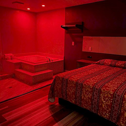 A room lit in red with a bed, a patterned bedspread, and an adjacent open spa bath area with tile steps.