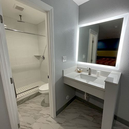 A modern bathroom with a white vanity, illuminated mirror, and a walk-in shower visible through an open door.