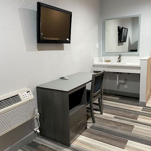 The image shows a room with a wall-mounted TV, small desk with a chair, a mirror above a sink, and a wall-mounted air conditioning unit.