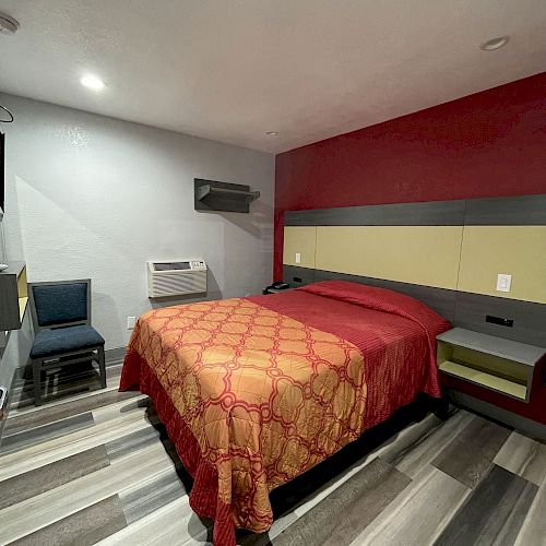 A neatly arranged bedroom with a bed, two chairs, and a small table against a wall. The bed has red and orange bedding, and the floor has striped wood.