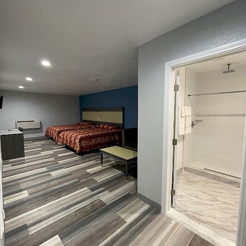 A modern hotel room with a double bed, TV, and dresser. A bathroom with a walk-in shower is visible to the right, all in a minimalist style.