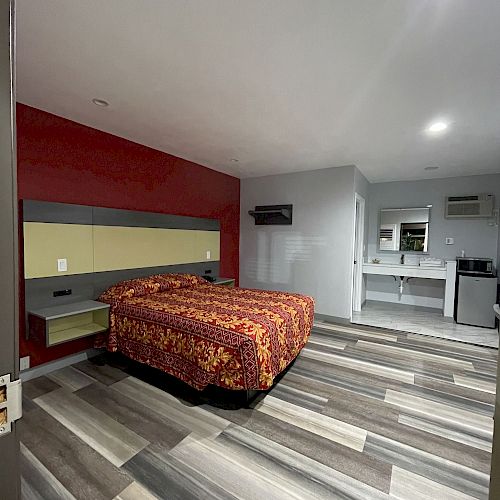 The image shows a modern bedroom with a patterned bedspread, wooden flooring, a partitioned bathroom area, a sink, and a small kitchenette.