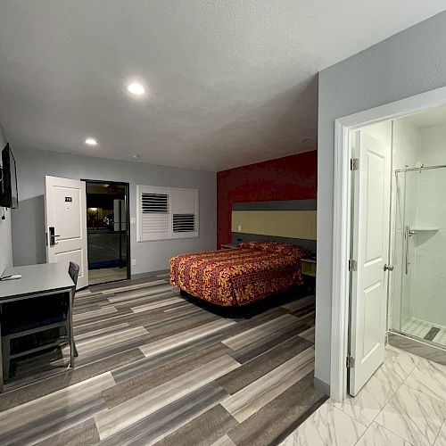 The image shows a hotel room with a bed, a desk, a chair, and a bathroom entrance. The bed has a patterned comforter, and the bathroom has a shower.