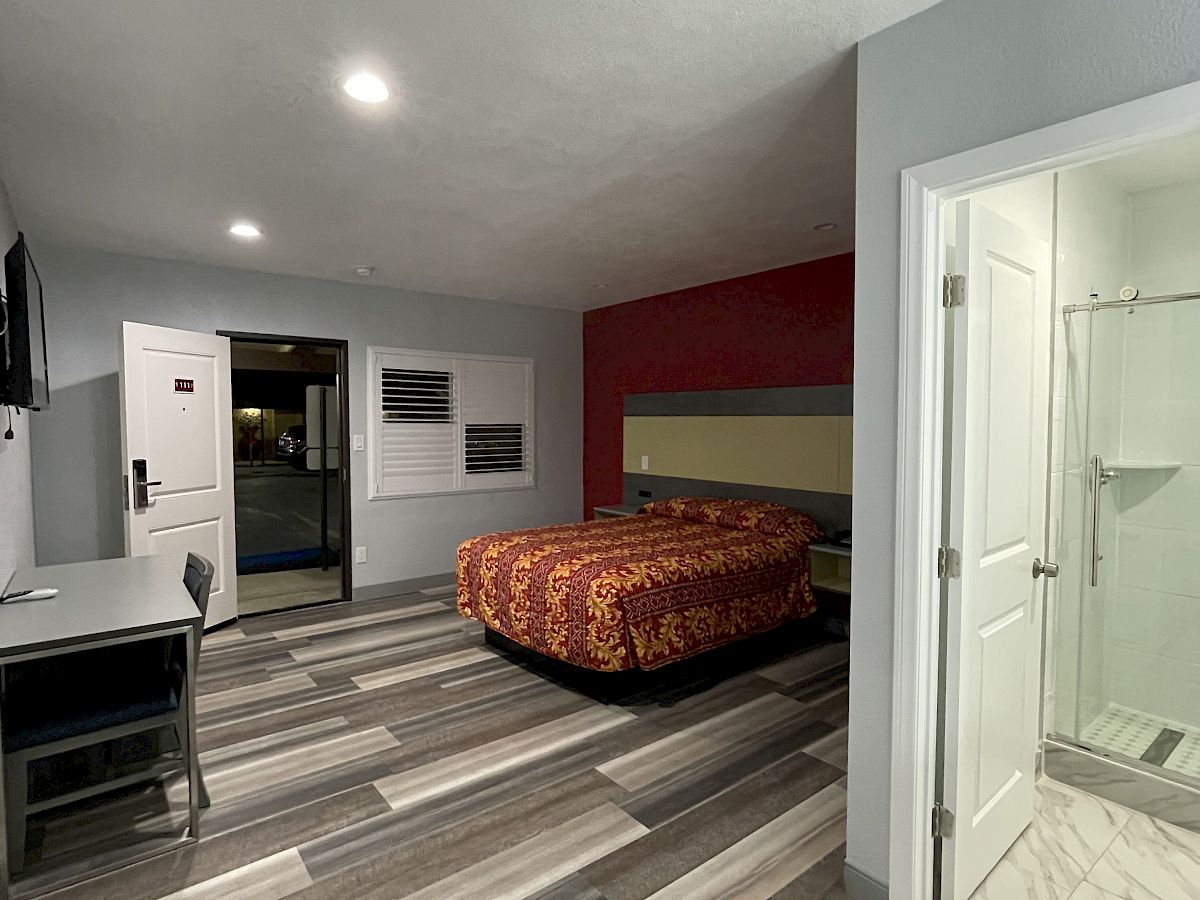 The image shows a modern hotel room with a bed, desk, TV, and an open door leading to the bathroom with a shower.