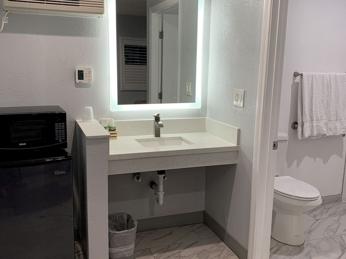 A bathroom with a sink, a mirror, a towel, and a toilet in a separate space. There's also a mini-fridge and an air conditioner on the wall.