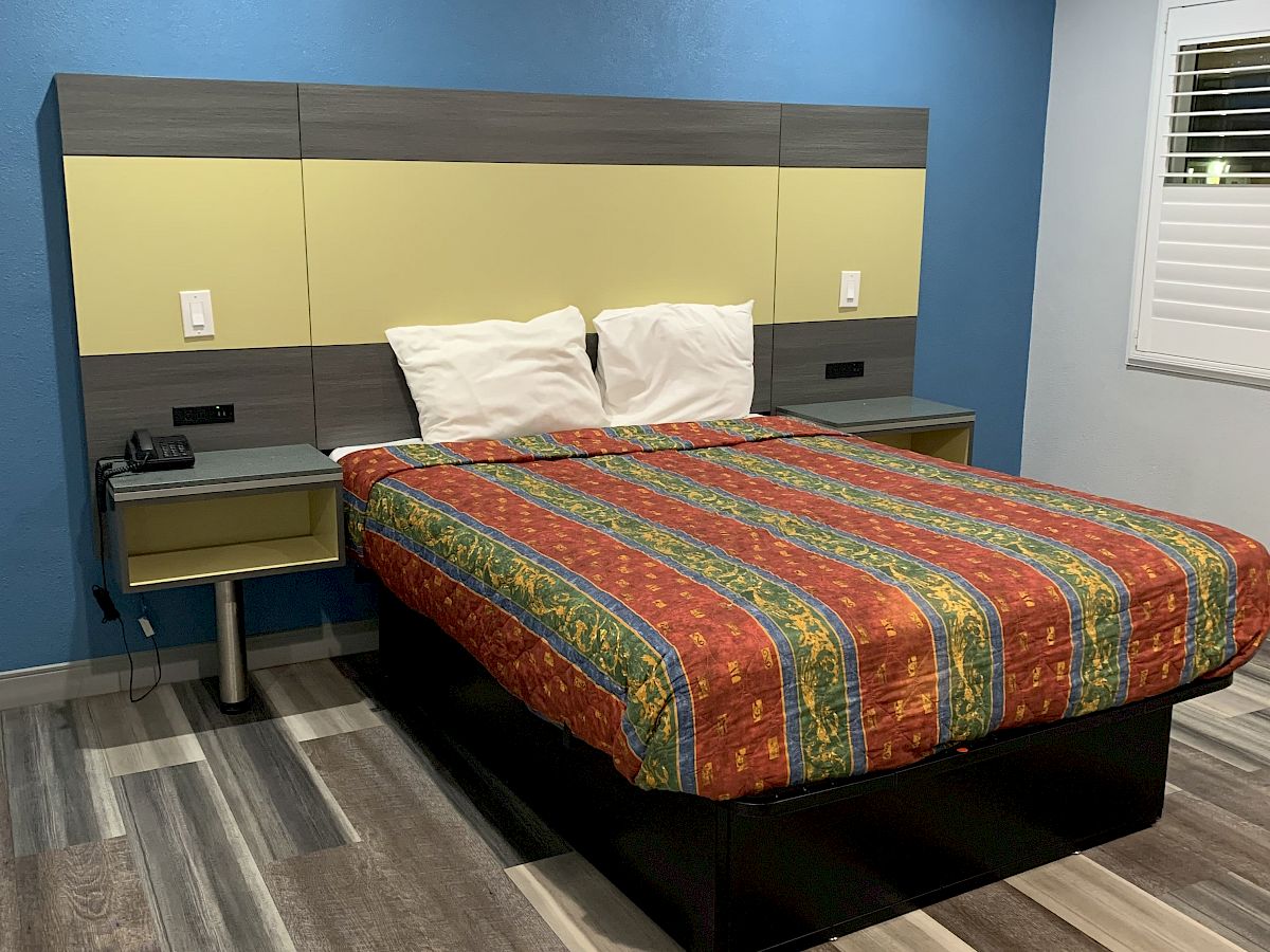 A modern bedroom with a colorful striped bedspread, blue wall, and wood-look flooring. The bed has a headboard with built-in nightstands.