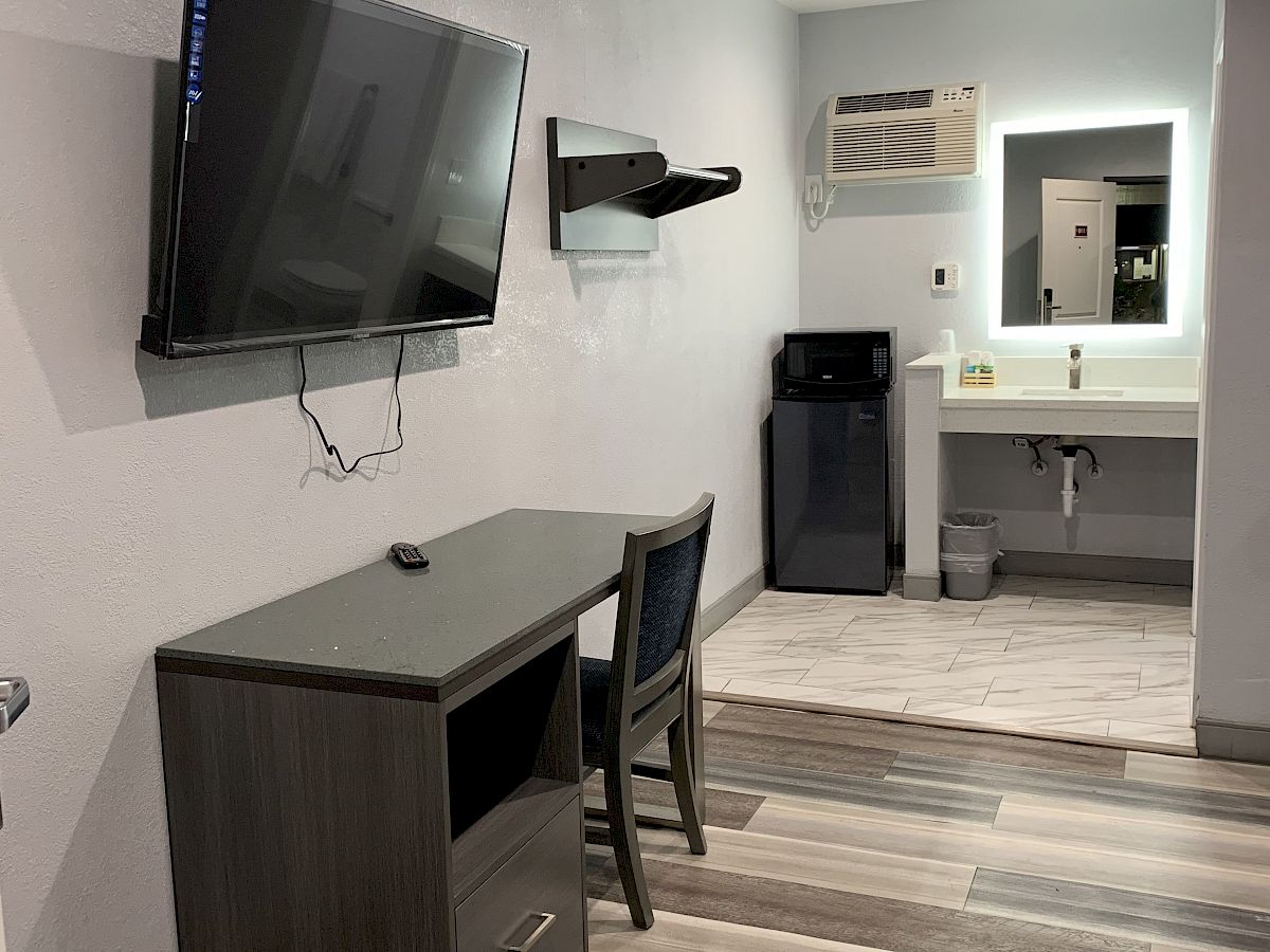 A modern room featuring a mounted TV, desk and chair, small refrigerator, sink area with mirror, and light wood-patterned flooring.