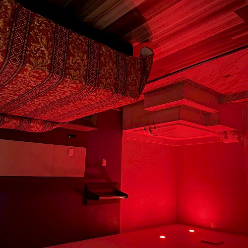 A hotel room with red lighting features a bed, patterned bedspread, jacuzzi-style bathtub, wooden flooring, and minimalist decor on the walls.