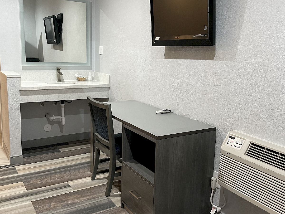 A small modern room with a desk, chair, wall-mounted TV, and air conditioner. There's also a sink area with a mirror in the background.