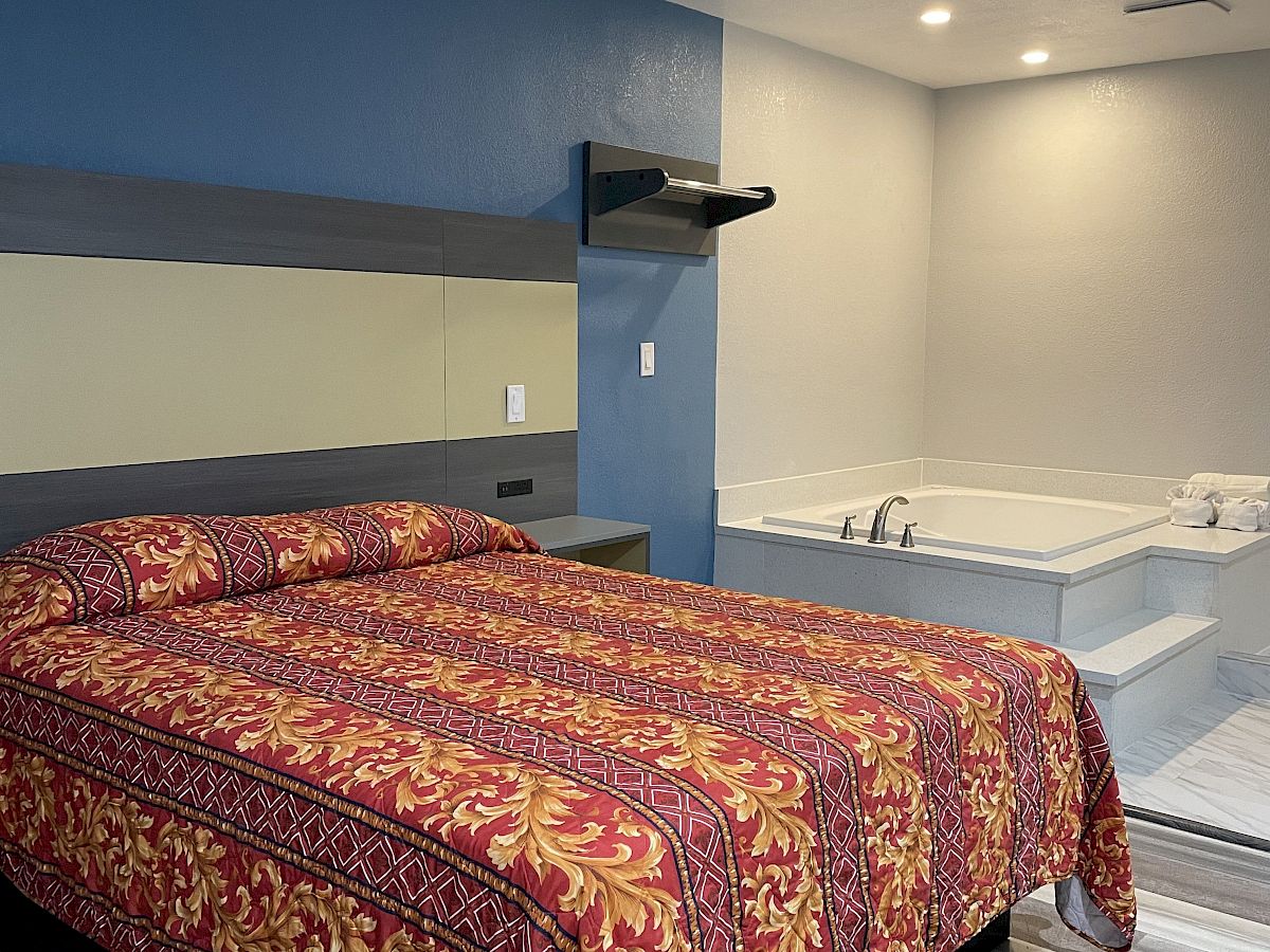 A bedroom with blue and gray walls, a bed with a patterned red blanket, a mirror, and a built-in bathtub in an adjacent area.