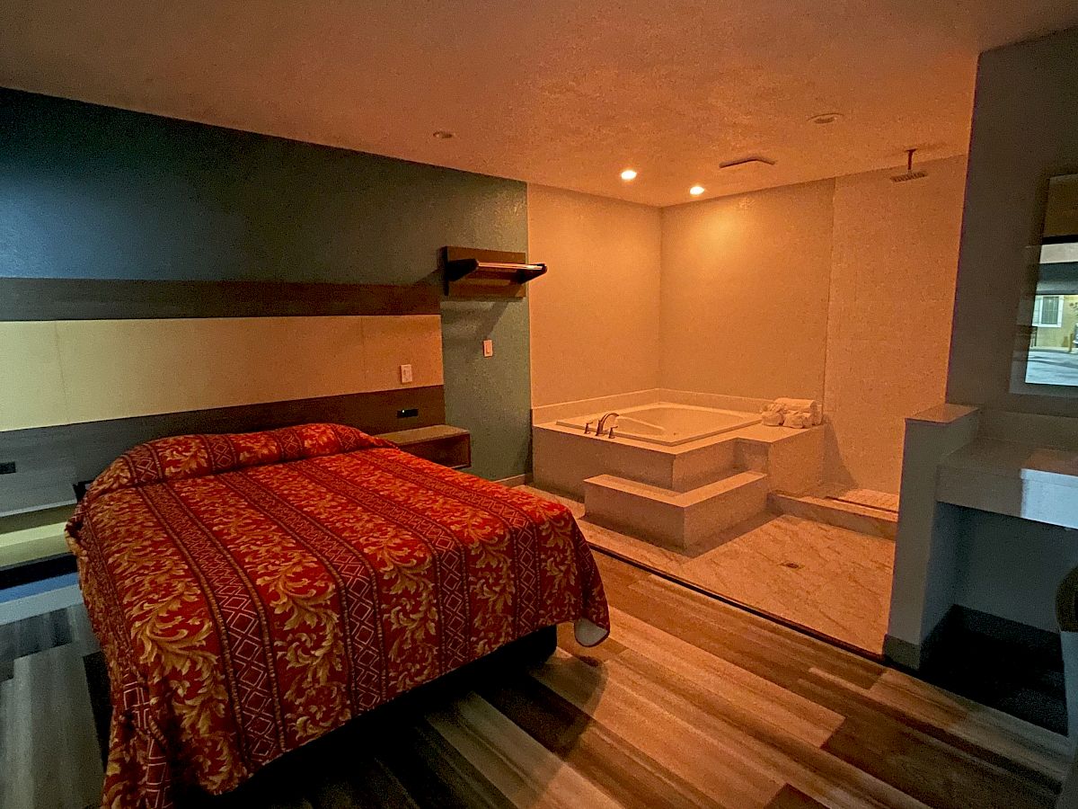 This image shows a bedroom with wood flooring, a bed with a red and gold bedspread, and a corner jacuzzi tub next to a small sink.