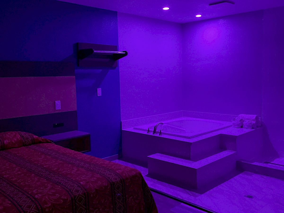 A room with purple lighting contains a bed, storage shelves, and a jacuzzi tub in the corner.