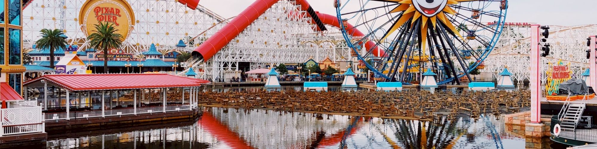 The image shows an amusement park featuring a large Ferris wheel with a recognizable face, roller coasters, and their reflections on a calm body of water.