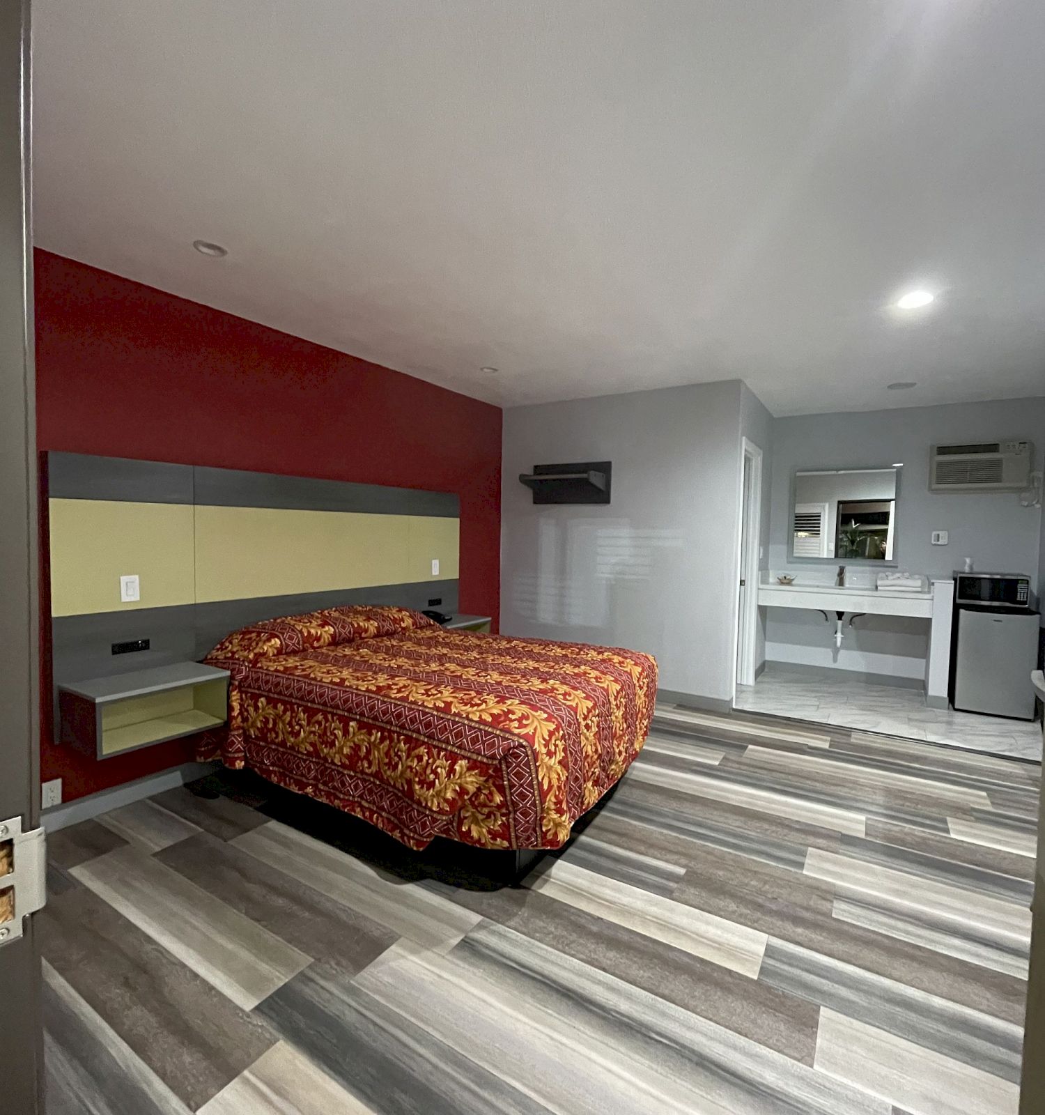 The image depicts a modern, sparsely furnished bedroom with a double bed adorned with a colorful bedspread, a sink, and a mirror in the background.