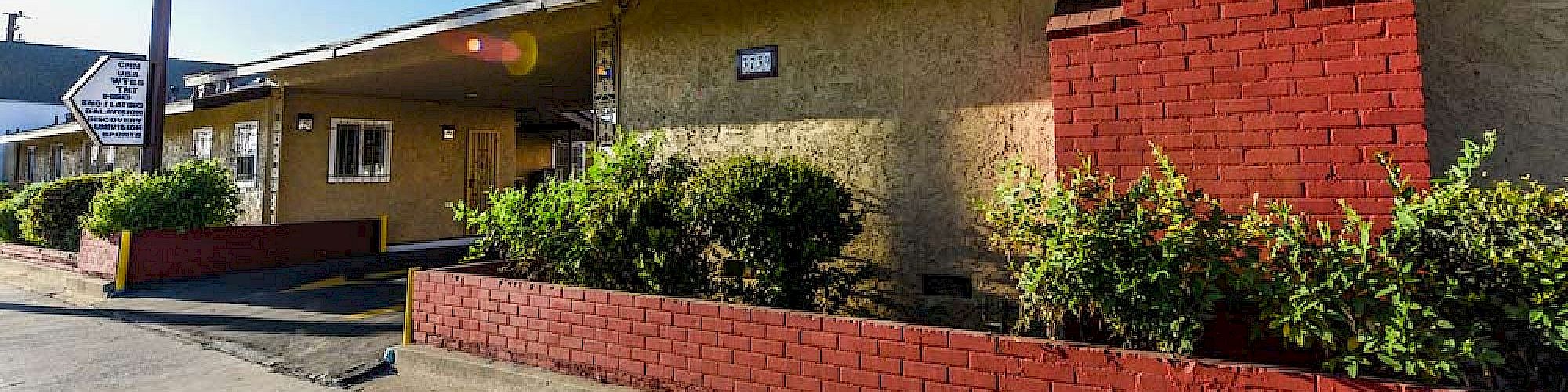 The image shows the exterior of a single-story motel with a "Motel" sign, beige walls, red brick chimney, and landscaped bushes in front.