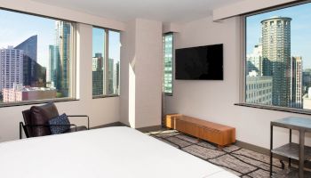 A modern hotel room with large windows showing a city skyline, a wall-mounted TV, a lounge chair, and a bed visible in the foreground.