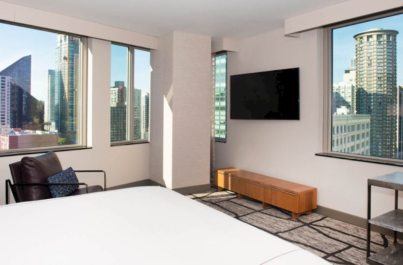 A modern hotel room with large windows showing a city skyline, a wall-mounted TV, a lounge chair, and a bed visible in the foreground.
