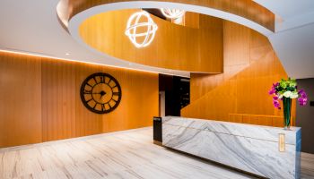 The image shows a modern lobby with a large clock on the wall, a marble reception desk, and wooden accents and curved lighting fixtures.