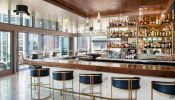 The image shows a modern bar with a sleek design, featuring high stools, a wooden counter, and shelves filled with various bottles of alcohol.