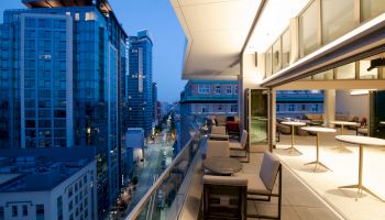 A balcony with chairs and tables overlooks a city street lined with modern buildings during twilight, with large windows offering an indoor view.