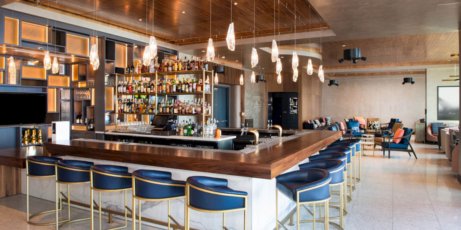 The image shows a modern bar with a wooden counter, blue cushioned stools, hanging lights, and a well-stocked shelf of liquor bottles in the background.
