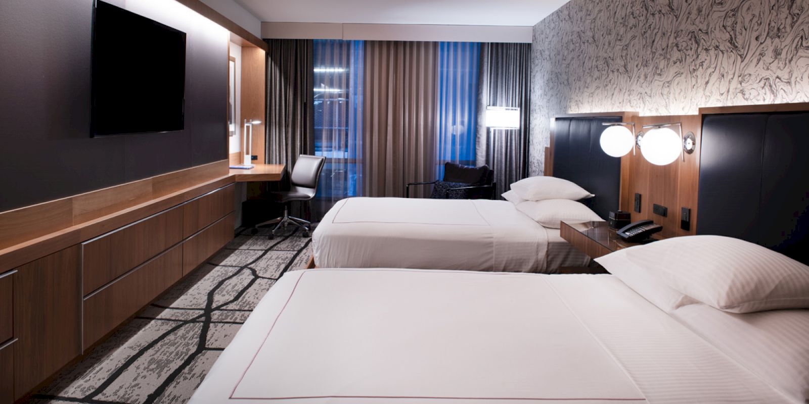 A modern hotel room featuring two beds, a flat-screen TV, a desk, a chair, and ambient lighting with a window covered by curtains in the background.