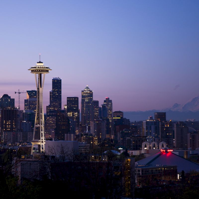 A city skyline at dusk with a prominent tower, tall buildings, and a distant mountain under a purple sky, showcasing an urban and natural landscape.