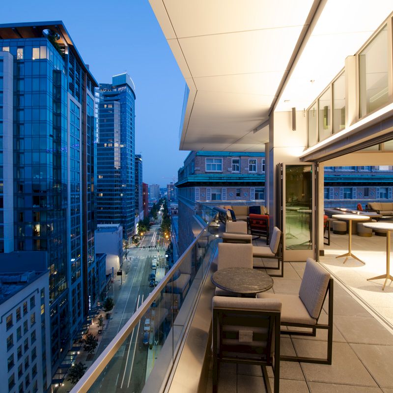 A modern rooftop terrace at dusk, featuring tables and chairs, overlooking a cityscape with tall buildings and a street below.