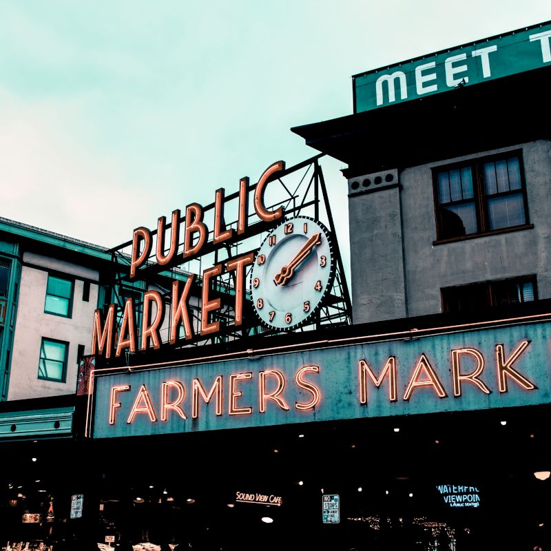 The image shows a public market with prominent signs saying "Public Market" and "Farmers Market," along with a clock and surrounding buildings.
