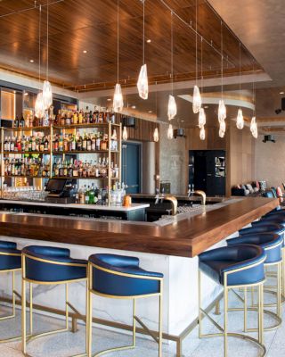 A modern bar with blue and gold bar stools, a well-stocked back wall of liquor, pendant lighting, and a lounge area in the background.