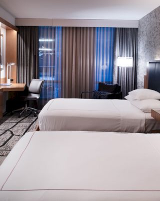 A modern hotel room with two double beds, a flat-screen TV, a desk and chair, bedside lamps, and patterned wallpaper, with large windows.